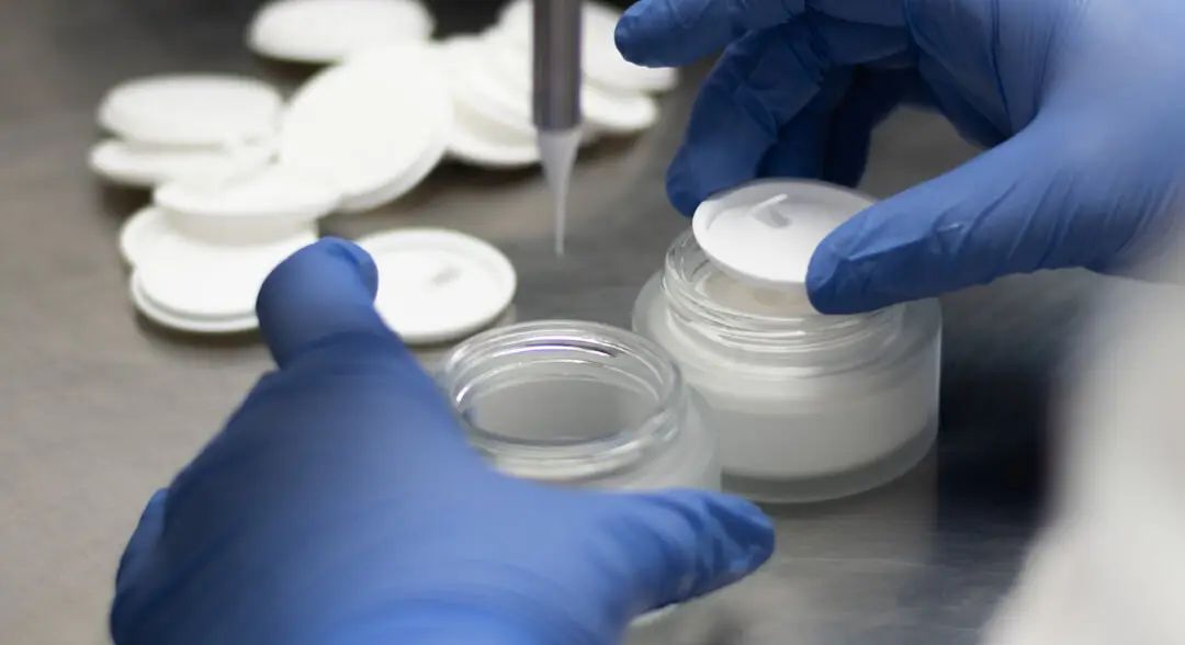 zwei Glastiege werden mit Deocreme gefüllt Person trägt blaue Handschuhe im Hintergrund liegen weiße Zwischendeckel